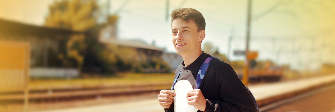 junger Mann mit Rucksack steht auf Bahnsteig und lächelt